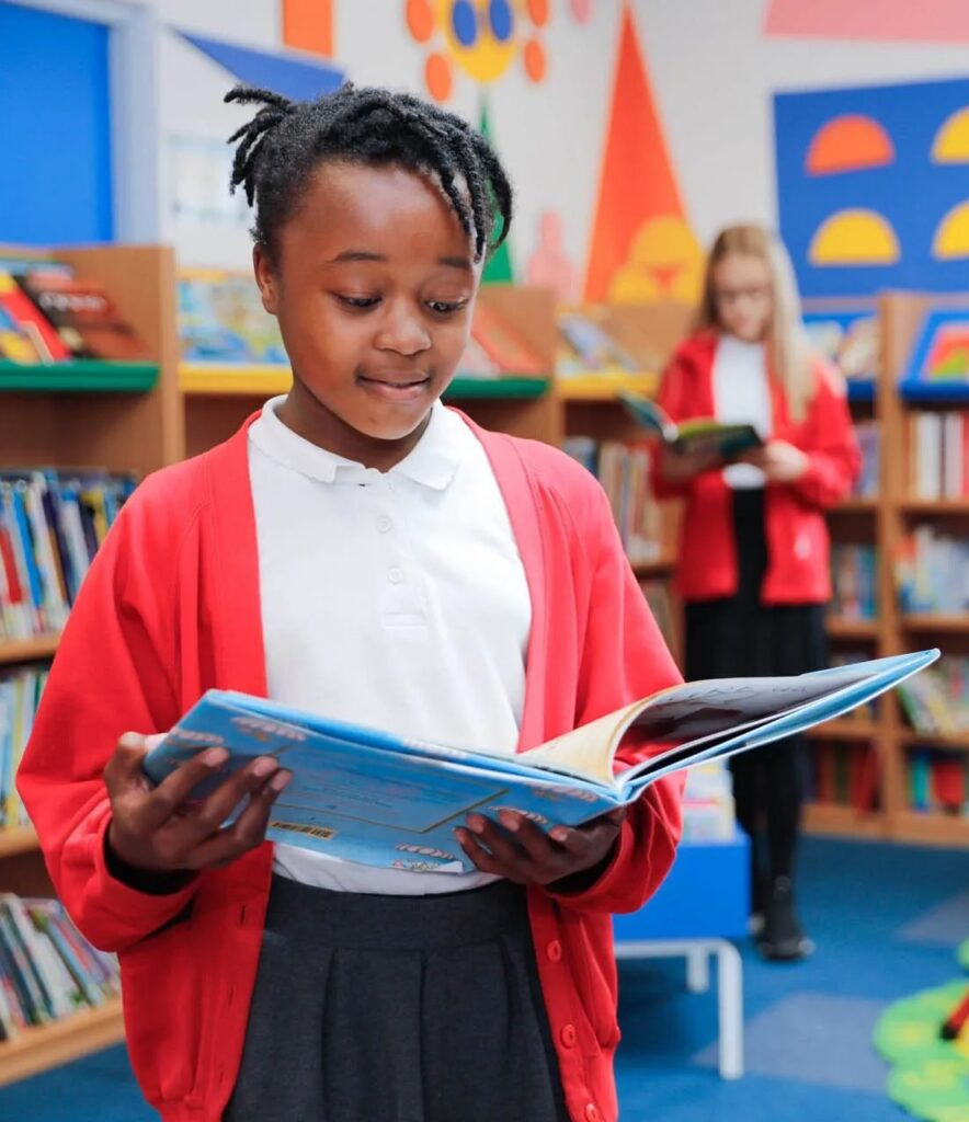 Young school girl in red and grey uniform, standing and reading a book in the library with another school girl in uniform reading another book.